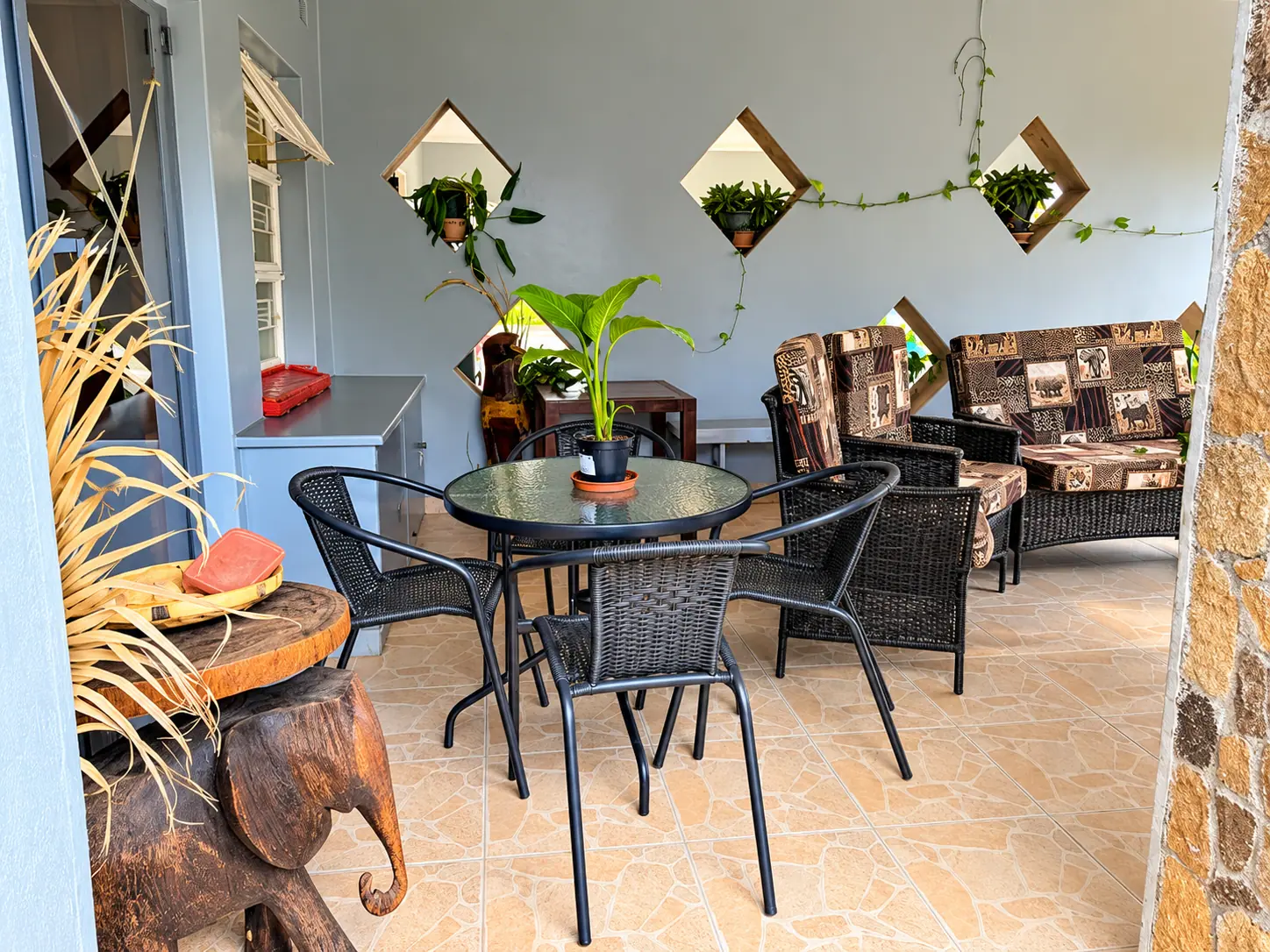 A round glass table with wicker chairs on the covered patio, beside a carved wooden elephant side table