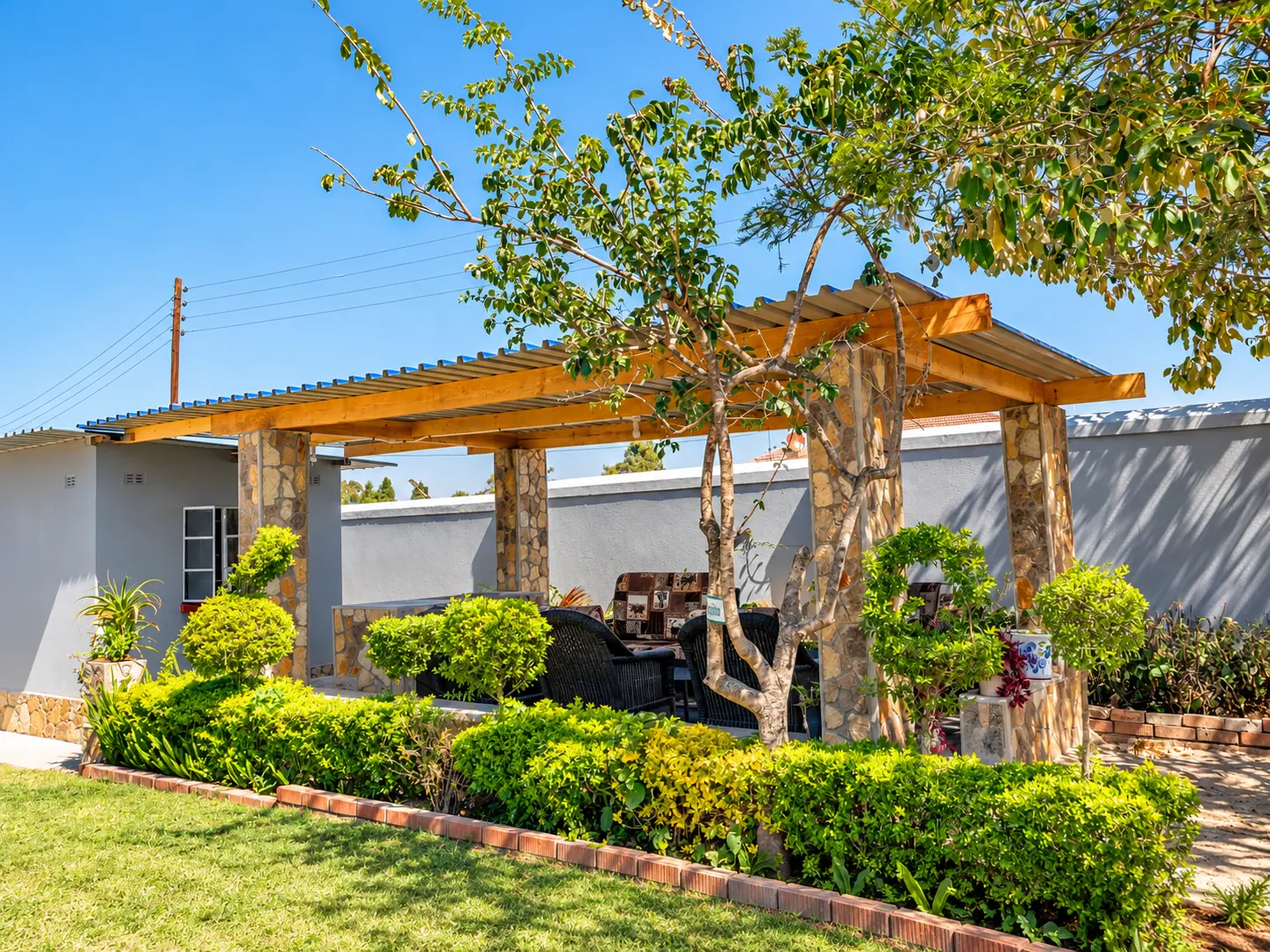 A timber pergola on stone pillars, framed by clipped hedges and a tree in the garden
