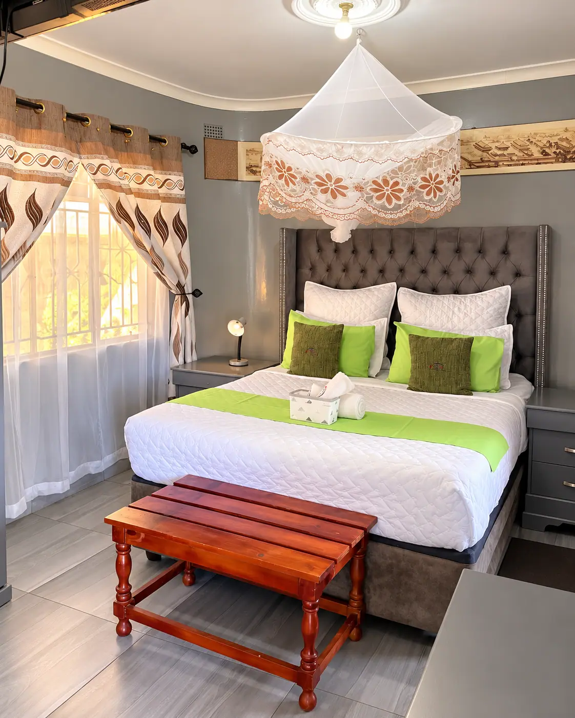 A double bed with a grey tufted headboard, green cushions and a lace-trimmed canopy net