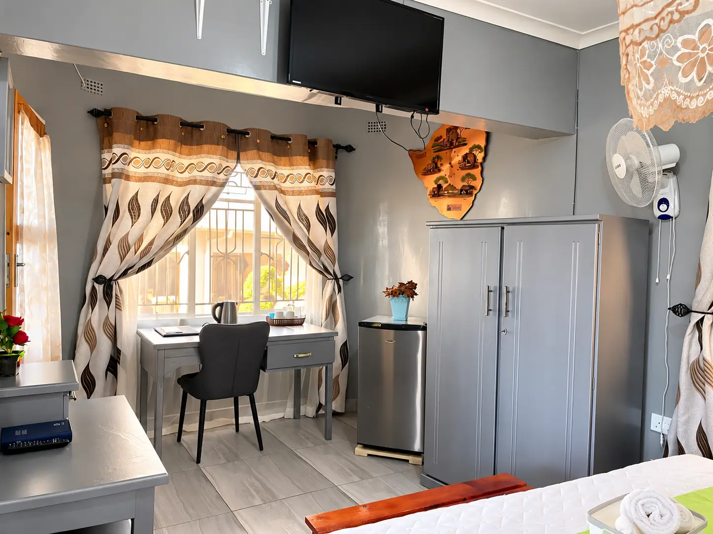 A writing desk by the window, with a bar fridge, wardrobe and wall-mounted TV