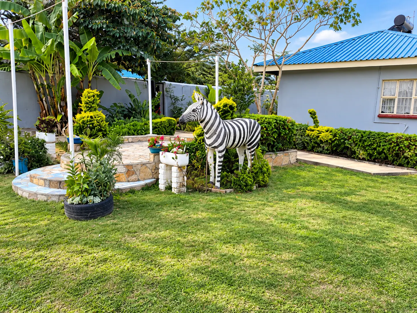 A zebra sculpture beside a round stone platform on the lawn, with a blue-roofed cottage behind